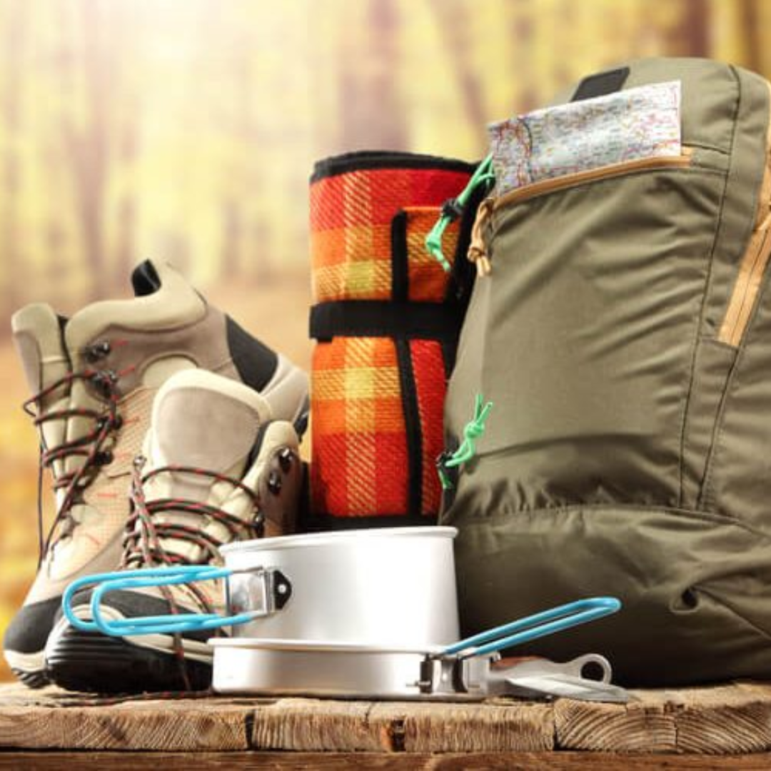 Hiking gear including boots, a backpack, and cooking equipment on a wooden surface with a blurred natural background.