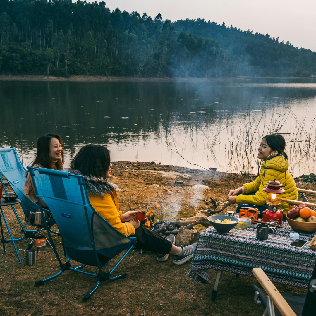 Three people sitting around a campfire by a lake, enjoying a picnic.