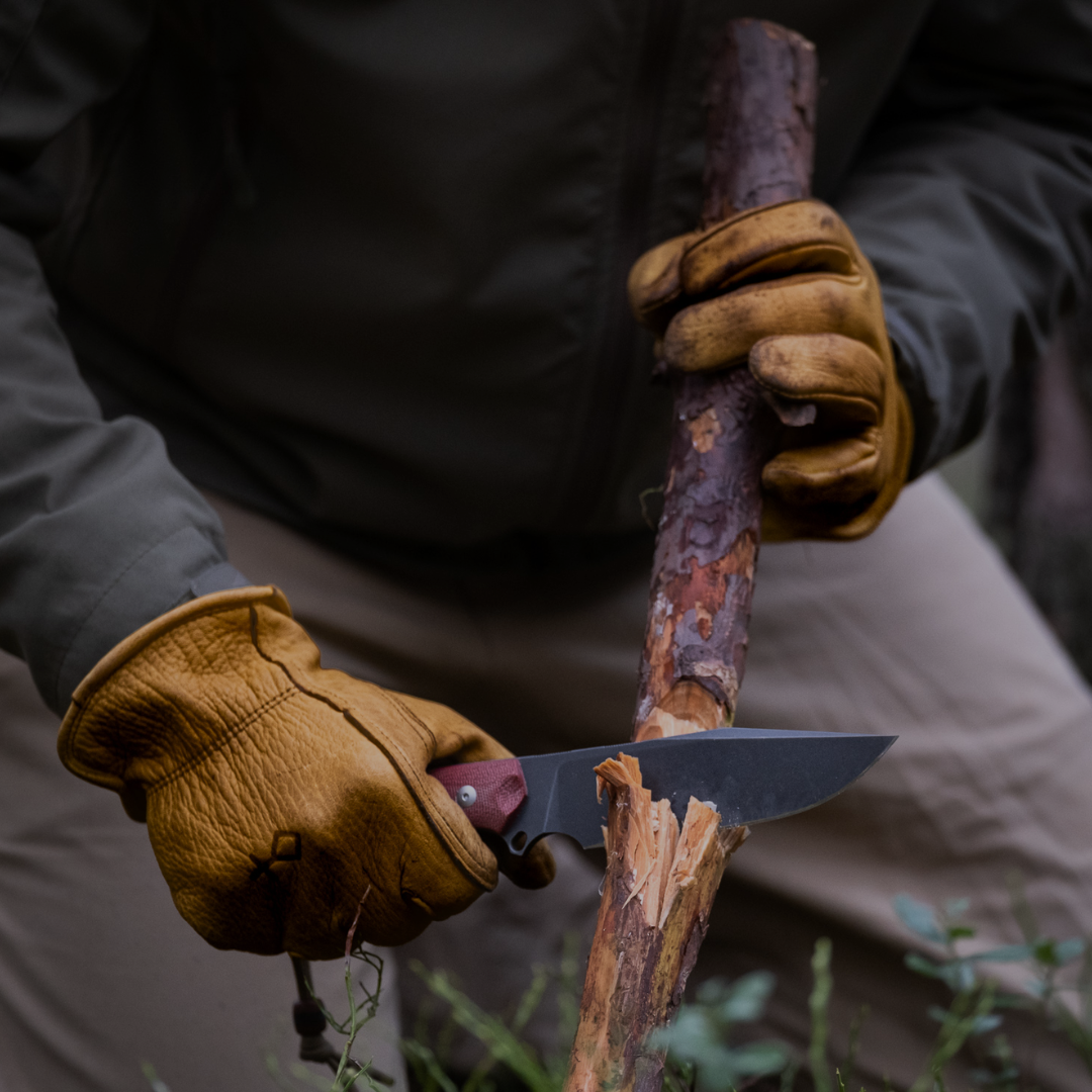 Person wearing brown gloves holding a knife and cutting wood with a natural background