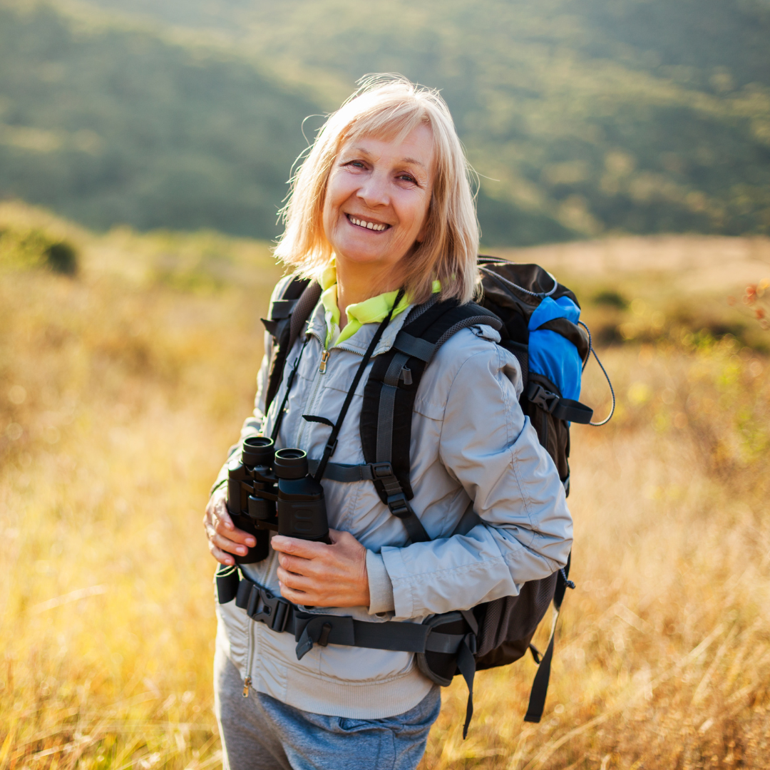 Woman with a backpack and binoculars standing in a field with mountains in the background