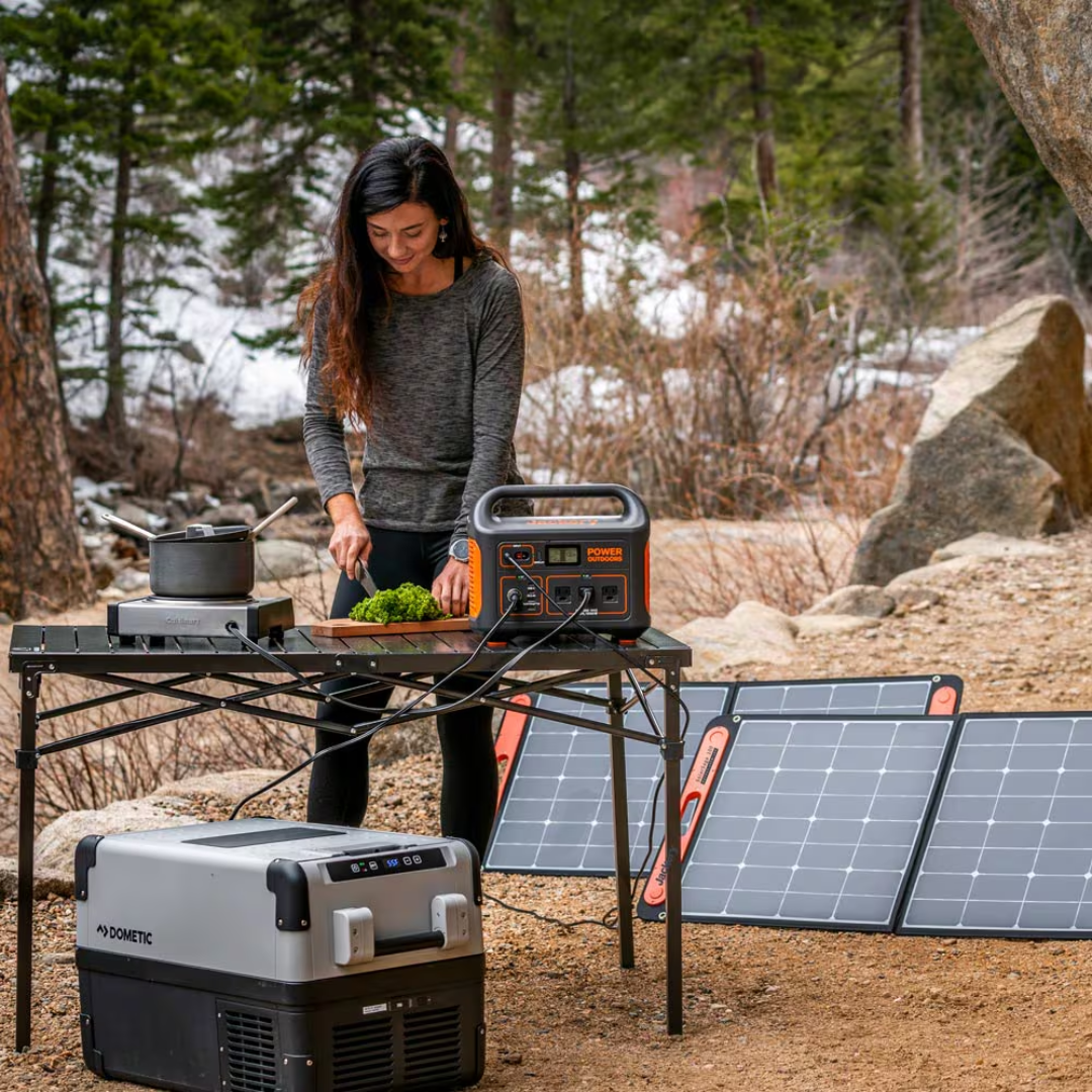 Person preparing food outdoors with a portable power station and solar panel in a forest setting.